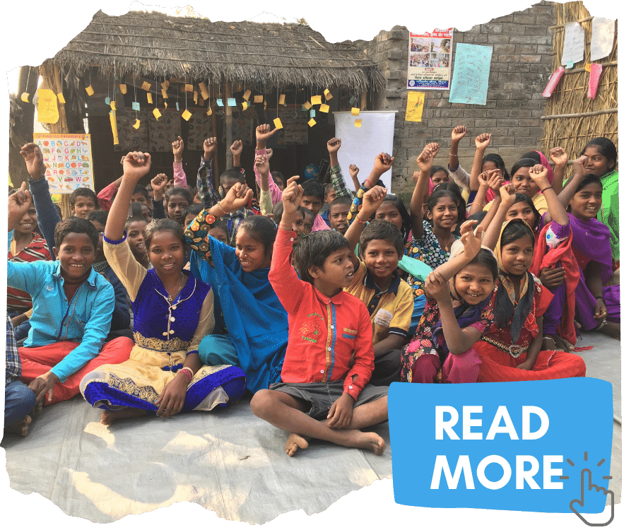 A group of Dalit children sat cross legged on the floor outside their classroom. They are all smiling and holding one arm in the air, making fists. You can click on the image to read more about how we tackle caste discrimination.