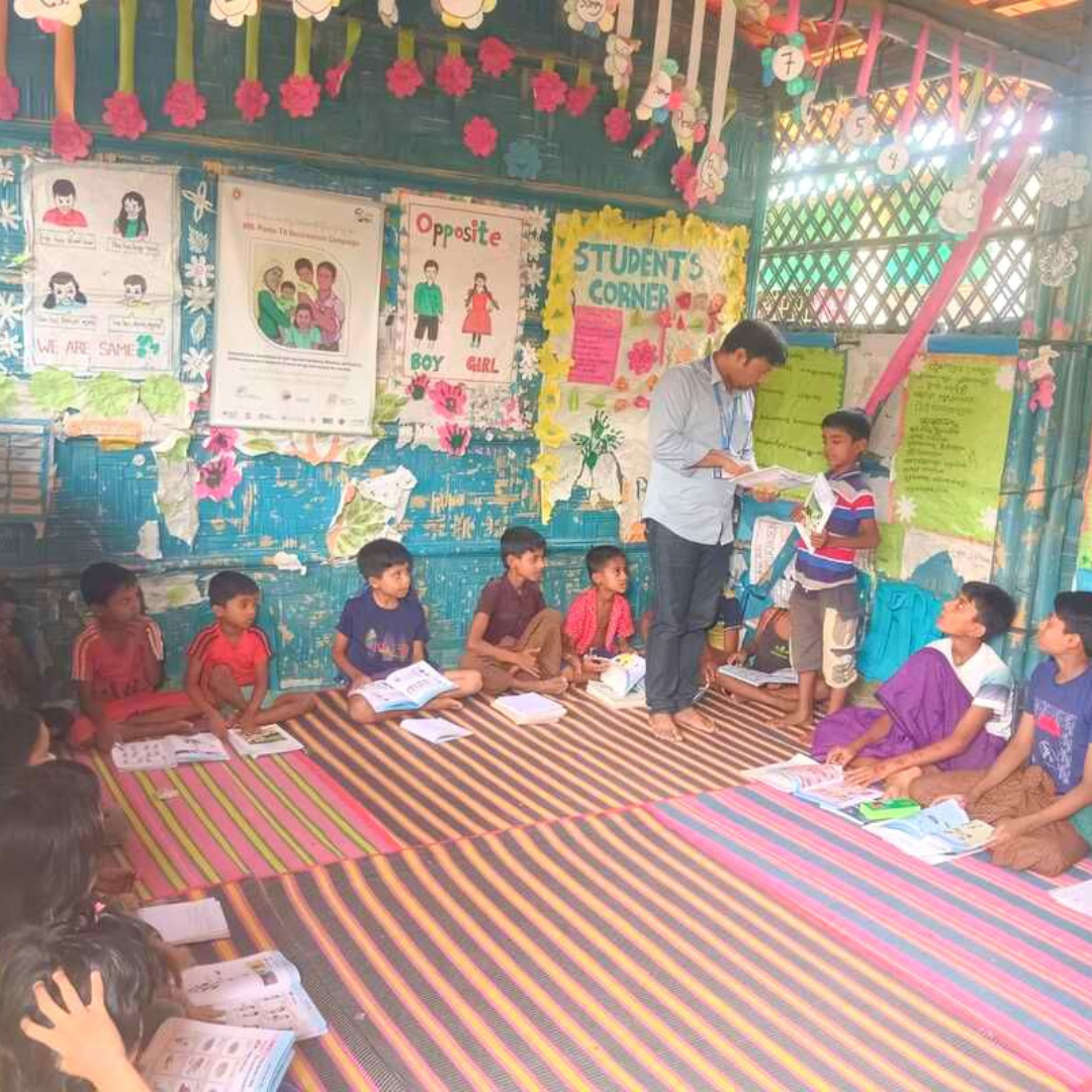 This is an image of Somorjit, a project officer in Bangladesh. He is kneeling behind two students in a colourful classroom and is smiling at the camera. Somorjit is wearing a maroon shirt and has his arms around the two students in front of him. The students are two boys who are also smiling and are sat behind a colourful table with books and stationary piled in front of them. You can click on the image to read the blog post. 