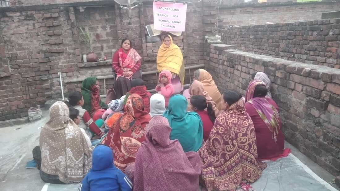 A group of Indian mothers pictured from behind taking part in a group counselling session run by two indian women sat at the front. They are sat outside on a mat, surrounded by brick walls 