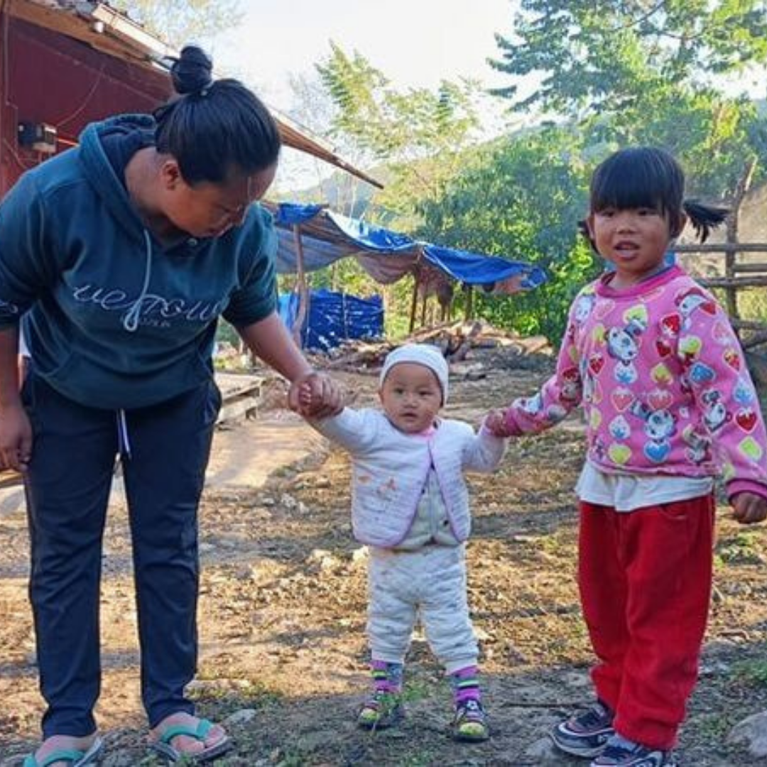 A toddler from Kachin State Myanmar is walking along holding hands with an older girl on her left and a woman on her right. They are wrapped in warm clothes as they live in a high altitude settlement for displaced people in Kachin State. 