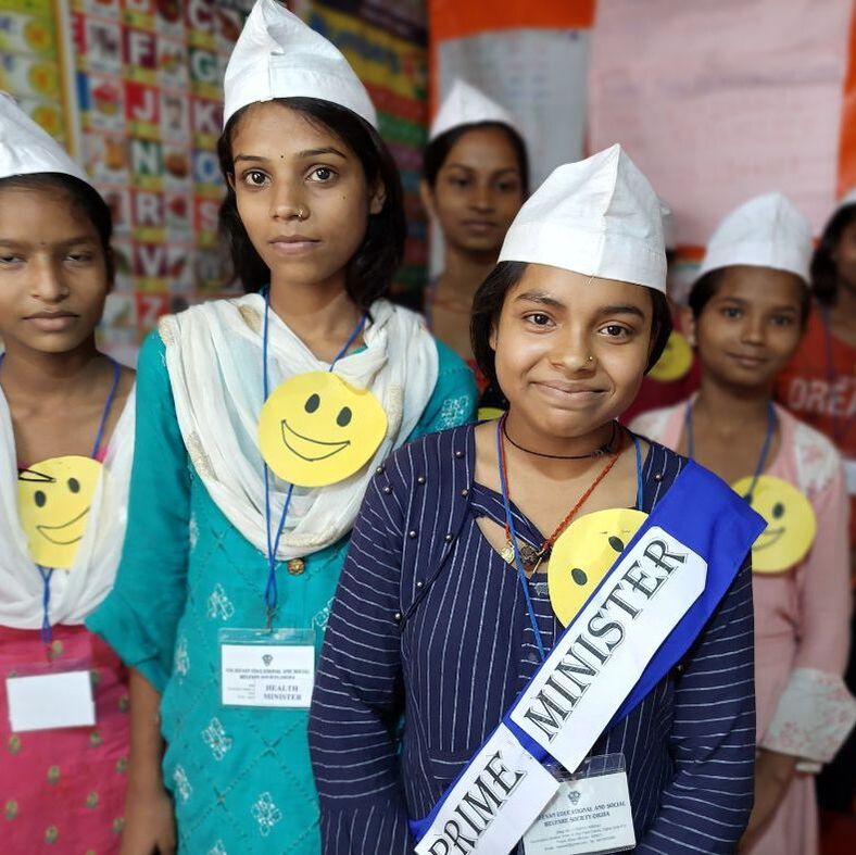 5 Indian girls are stood smiling at the camera wearing white hats. One at the front has a sash on which reads 'prime minister'. They are members of one of the Child Parliaments we support in India