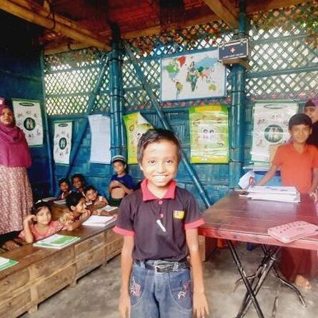 Abdul, a small boy wearing a black tshirt is smiling at the camera in his classroom. He has a big grin.