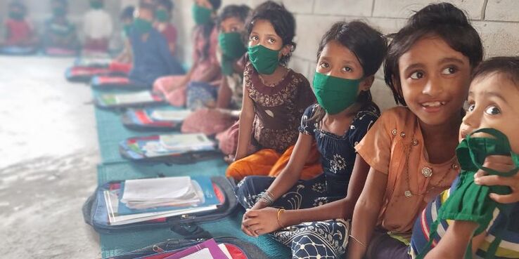 Four young Rohingya refugee girls are sat in their classroom on the floor against the wall. All but one are wearing matching green cloth face masks. They have new bags and books on the mat in front of them.