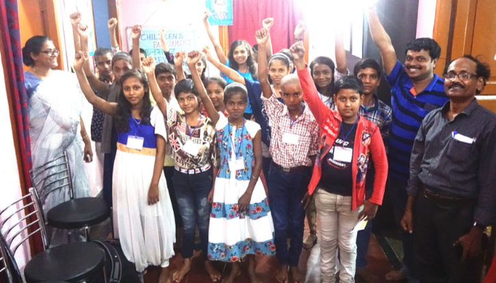 Indian child parliament members standing together in a crowded room with one arm in the air held in a fist. They are smiling. Three adults stand beside them.