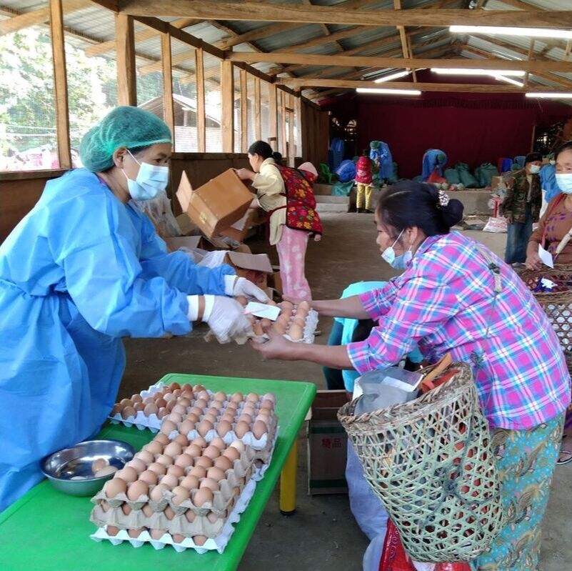 Humanitarian relief in Kachin - a woman wearing PPE is handing out a tray of eggs to a Kachin woman carrying a large basket