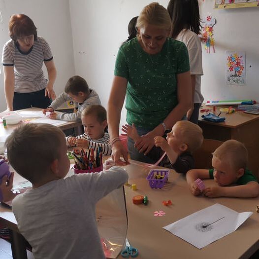 A woman in a green shirt is with two other adults, supervising a group of young Ukrainian refugee children. They are drawing and colouring in at a table.