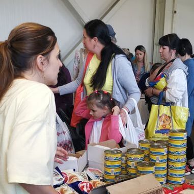 A woman is helping at a drop in centre in Romania behind a table filled with tinned goods and other food. Queues of Ukrainian refugees are waiting to collect items. 