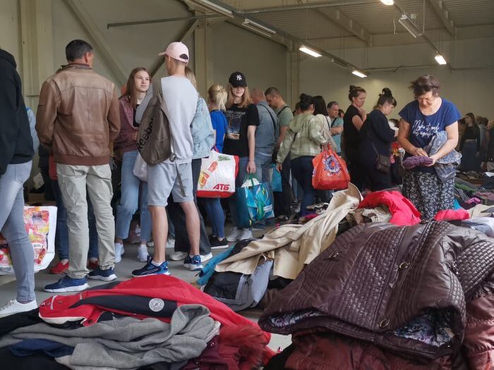 A pile of clothes, ready to collect is waiting on one side of a large room where refugees are queuing up with bags ready to collect items