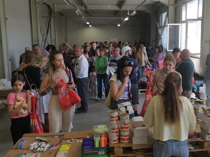 A large room is filled with people waiting, lining up to collect food tins from a table at one end of the room