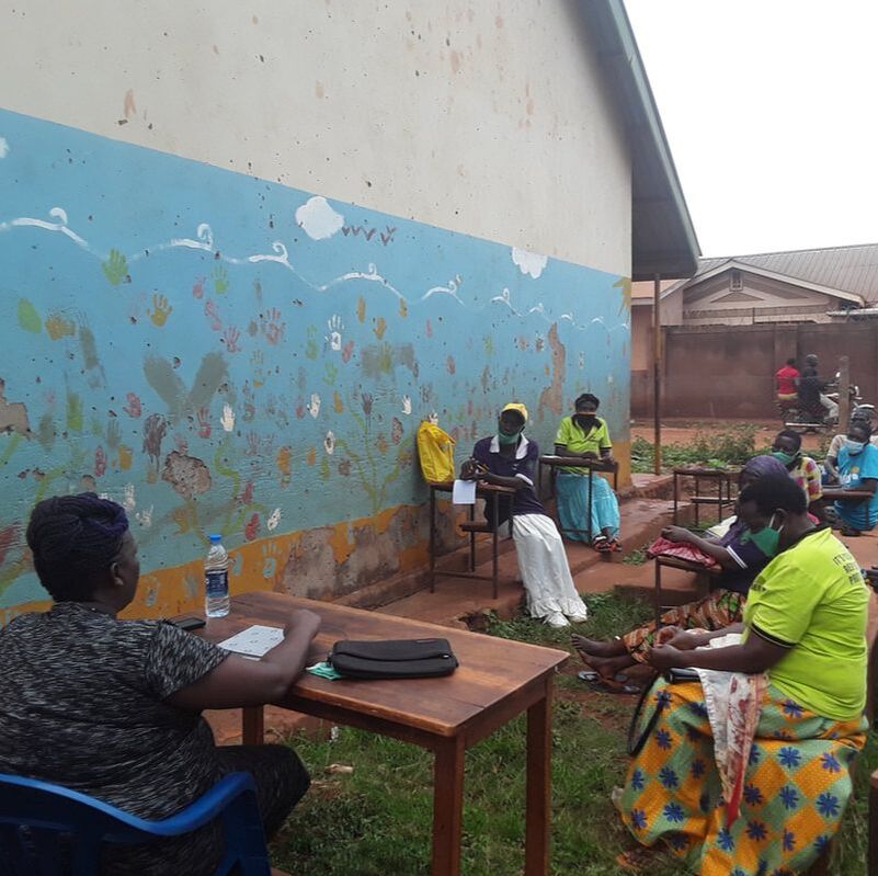 An outdoor workshop taking place in Masese II, participants are sat on chairs outside a painted walled building, listening to a woman speak at a table in front of them 