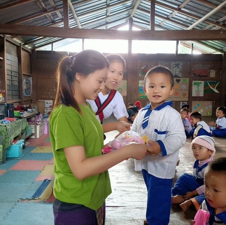 Image shows a young Kachin girl receiving a parcel from a woman  with a new raincoat and pair of rubber shoes. She is similing and they are both inside a classroom building with coloured drawings pinned up on the walls. 