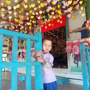 A young Kachin boy is peering round a pillar on the verandah of his colourful preschool classroom