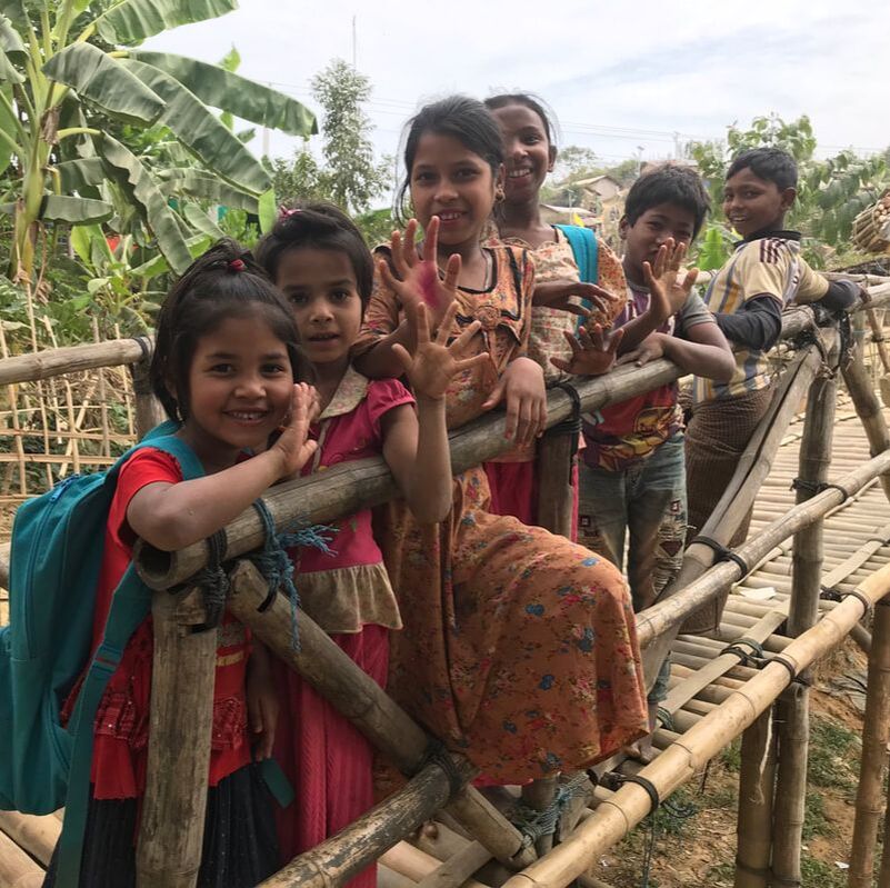Six young rohingya children are stood on a bamboo bridge, holding onto the railings and waving 