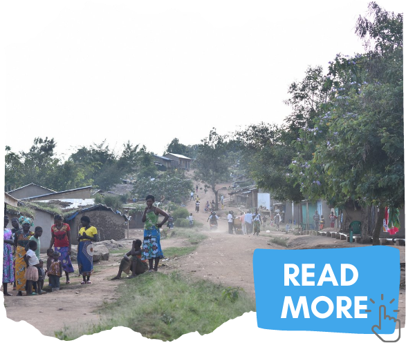 A dusty street in Kyaka II settlement. There are makeshift houses and shelters lining the road and lots of green trees. Congolese refugees are gathered in small groups along the side of the road. You can click on the image to read more about the issues is Kyaka II