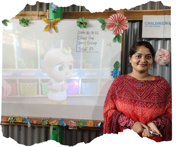 Ritu, a 27 year old Bangladeshi woman is stood in front of a projector screen showing cartoons in a corrugated walled classroom. She is smiling. 