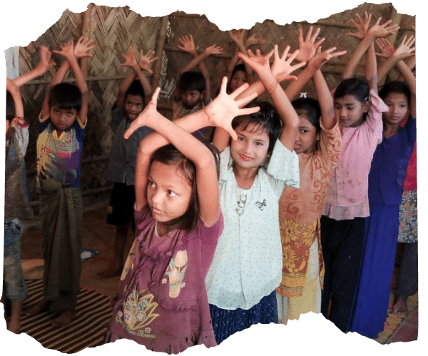 A group of young Rohingya refugee children are standing in two lines in their classroom with their hands above their heads, they are all smiling.