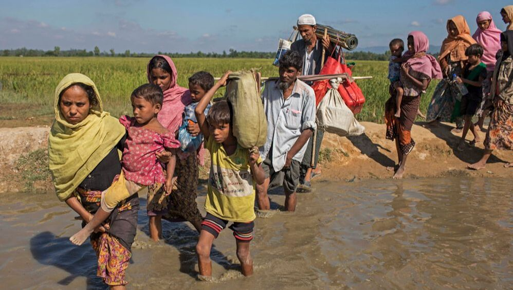 A line of Rohingya refugees laden down with belongings are crossing a stream. 
