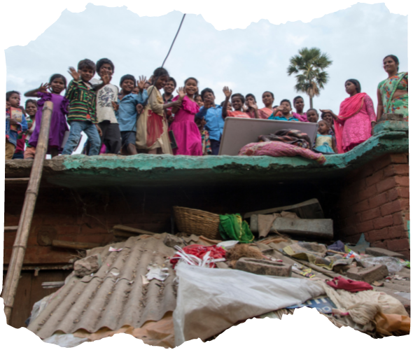 A group of Dalit children waving from a rooftop in a slum community. This is a rooftop school in a very crowed slum area. They are all smiling and you can see the ladder where they all climbed up. Their teachers are with them. The wall under the rooftop is stacked with rubbish. 
