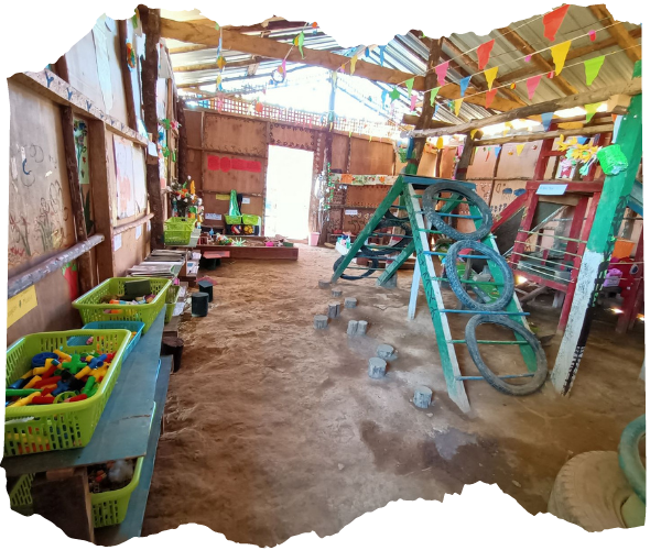 Play area constructed in an outhouse with homemade climbing frame, boxes of colourful toys and learning materials and brightly coloured bunting hanging all around the ceiling. 