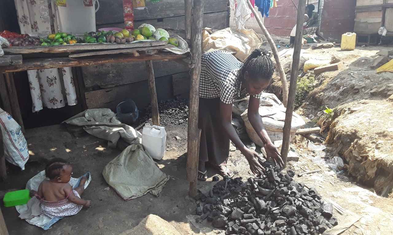 Scovia, a Ugandan woman is piling up pieces of charcoal in front of her stall in Masese I slum in Uganda. Sticks hold up the shelter where a wooden table holds vegetables. A small baby is sat on a mat in front of the stall. 