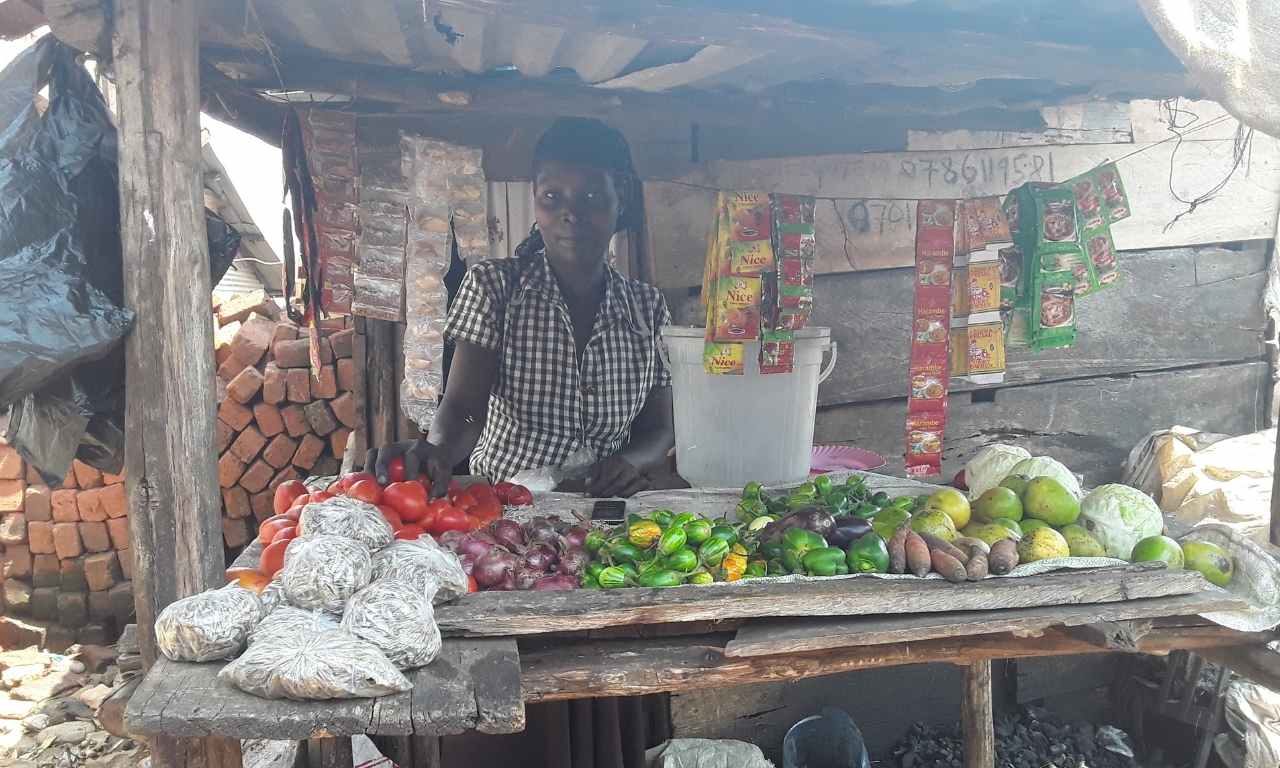 Scovia, a young Ugandan woman is standing behind a small stall with fruits and vegetables laid in front and small packets hanging on a string behind her