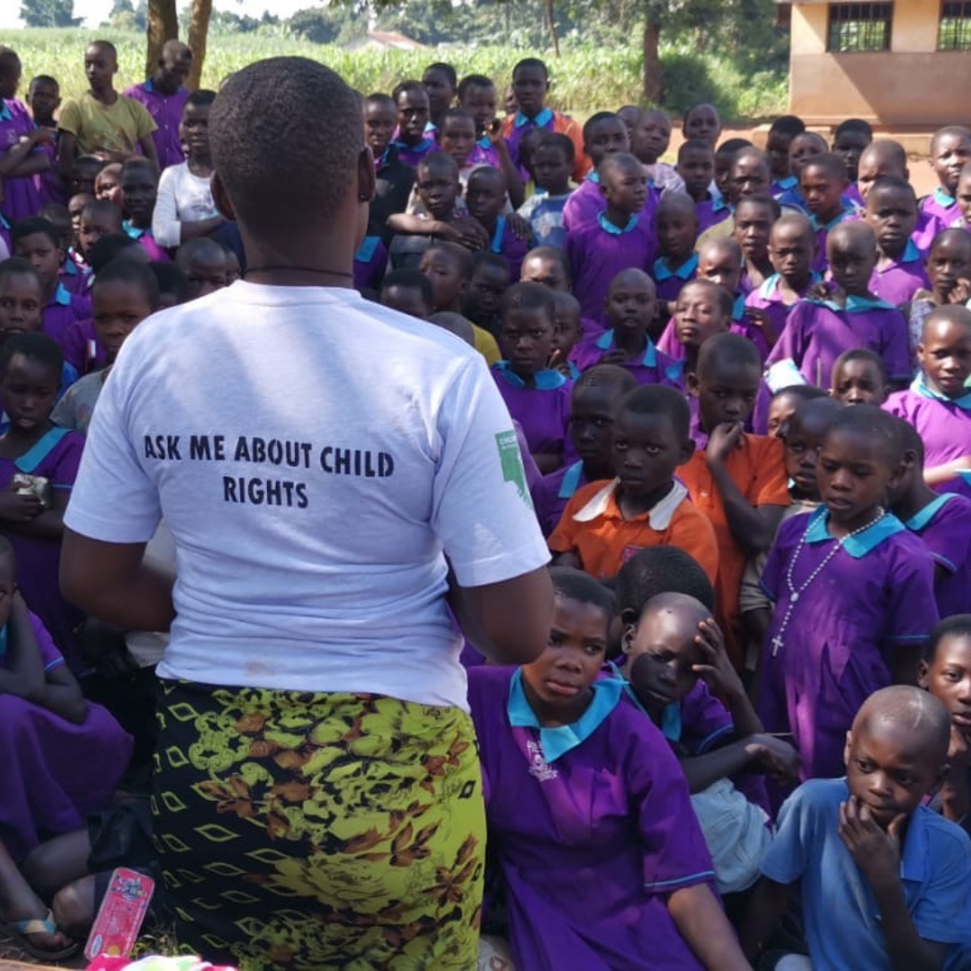 A Ugandan girl of about 17 stands with her back to the camera in front of a large crowd of younger children all in purple school uniform. She is giving a presentation to them and wearing a white T-shirt which on the back says 'Ask me about Child Rights'. 