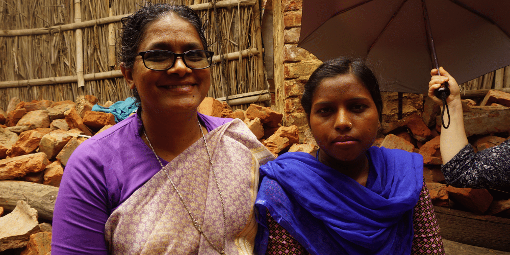 Image shows Sister Veena, an Indian woman wearing a purple tshirt and patterned purple sari / dress. She has dark brown hair and black rimmed glasses and is smiling at the camera. She is standing next to another woman wearing purple who is also looking at the camera, she is not smiling. 