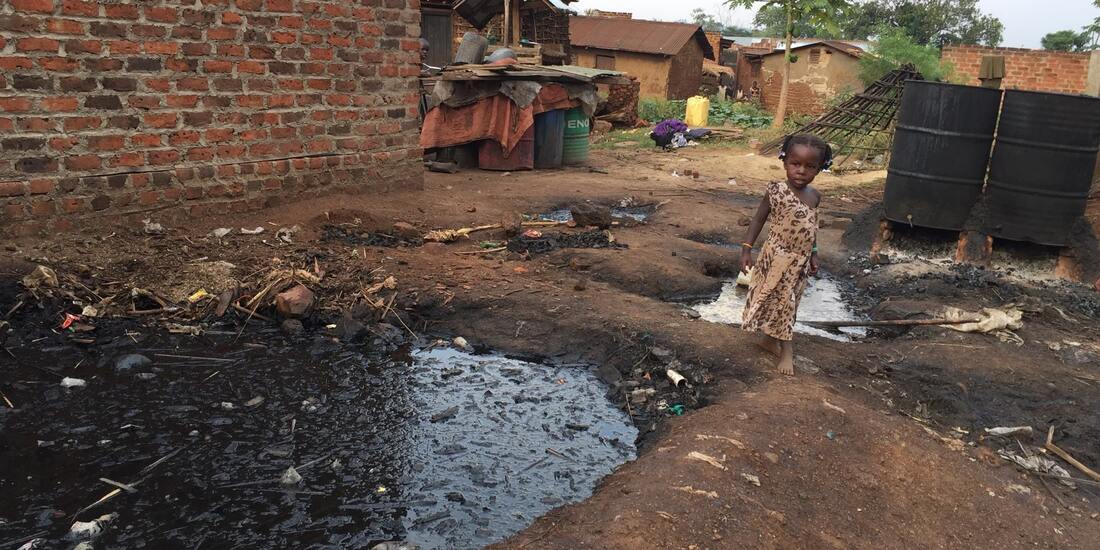 A Ugandan girl of about five years old walks through a muddy slum area which has blackened barrels used for brewing illegal alcohol. She is walking past a large pit of sludge which is the run off from this process. 