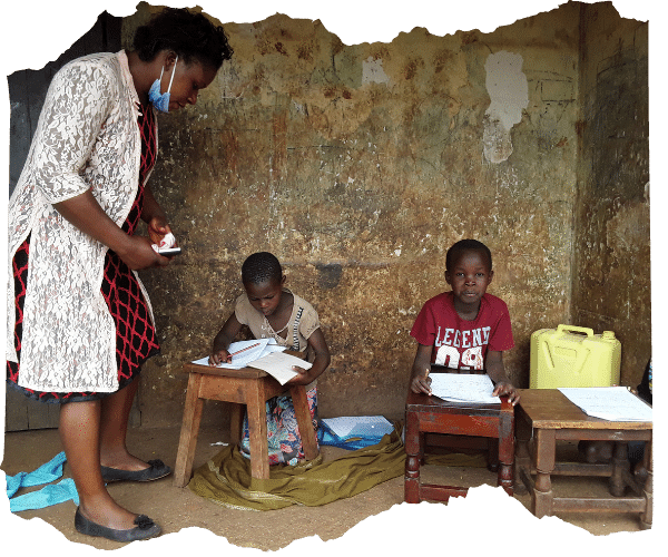 Ugandan teacher visiting two young children at home to deliver homework packs and check in on them. The two children are sat at little stools doing work as the teacher stands over them.