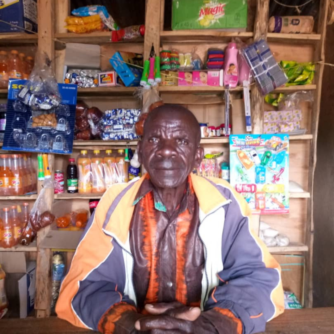 An older Ugandan man, Solomon with a serious face looking directly at the camera. He is wearing and orange and blue baggy jacket and is sat in front of his stall. Behind him are shelves filled with various goods - vegetable oils, snacks and a few other food items and household supplies. Solomon has his hands held in front of him and they are resting on a wooden desk in front of him. You can click on the image to read the blog.