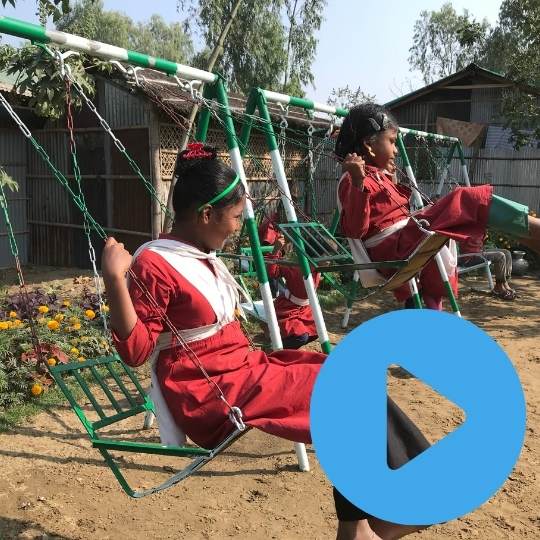 Two Bangladeshi girls in red school uniform swinging happily on metal swingsets in the garden of their community school in Bangladesh. Click on the image to watch the video.