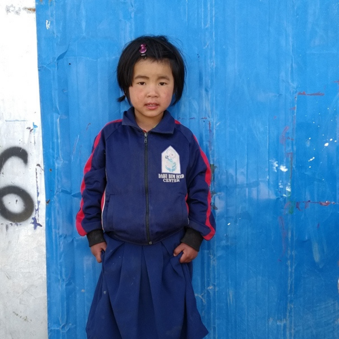Bawk Kai Mai, a young Kachin girl, aged five standing in front of a blue wall. She is wearing a navy blue jacket and skirt with some sort of logo on her chest and red stripes down her arms. She is looking at the camera with a straight face. She has black hair, clipped back on one side with a pink hair clip. 