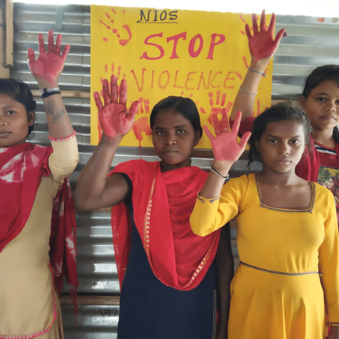 Four teenage Indian women standing in front of a wall made of corrugated metal. They are looking at the camera with serious faces and holding up their right hands. Their palms are painted red. A yellow poster on the wall behind them reads 'Stop Violence' with red hand prints around the edge. You can click on the image to read the blog.  