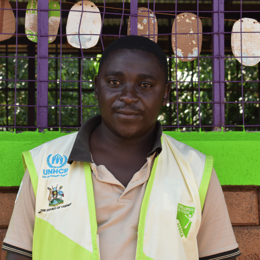 This is an image of Henry, a programme manager at Children on the Edge Africa.  Henry is standing in front of a new school building which has bright green walls and purple fencing for windows. Henry a black man with short hair. He is looking at the camera and smiling. He is wearing a hi viz vest with a Children on the Edge and UNHCR logo on it. You can click on the image to read the blog post. 
