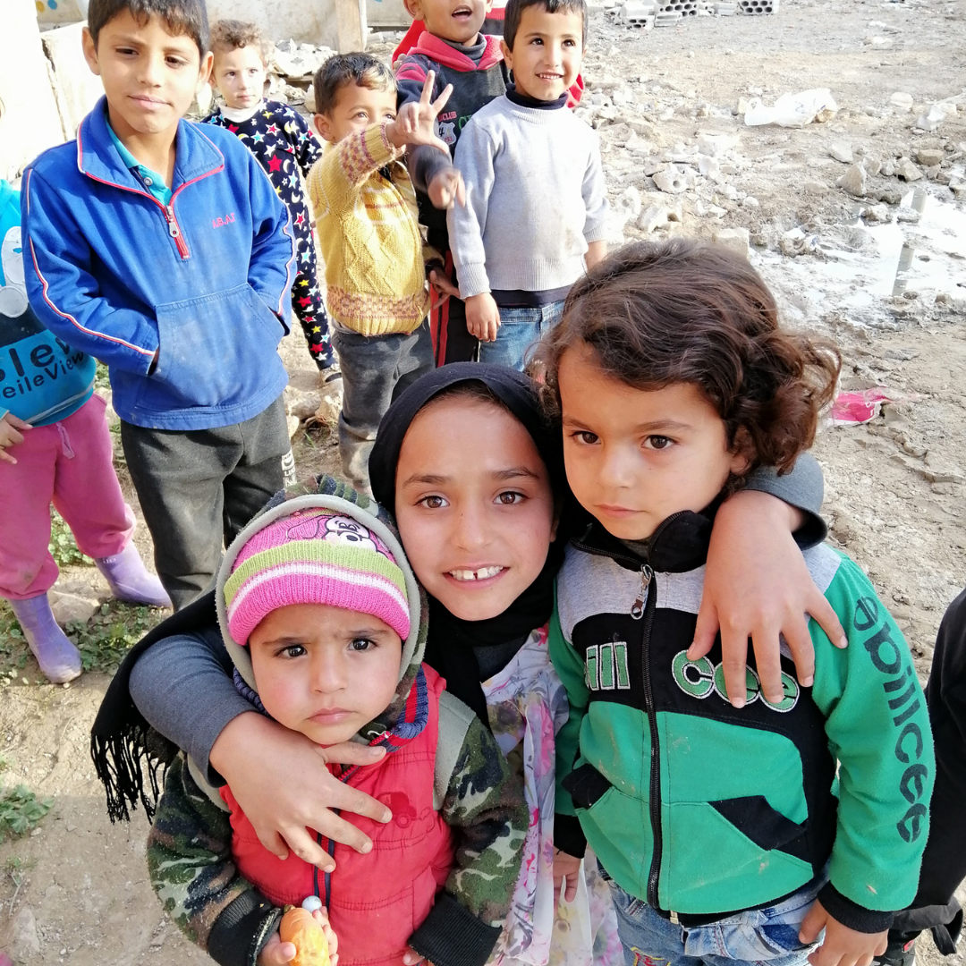 Hoda, a young Syrian girl wearing a black headscarf. She has her arms round two younger siblings, one on each side. Hoda is smiling. Behind her you can see five other children, young boys. They are standing on a muddy road. You can click on the image to read Hoda's story