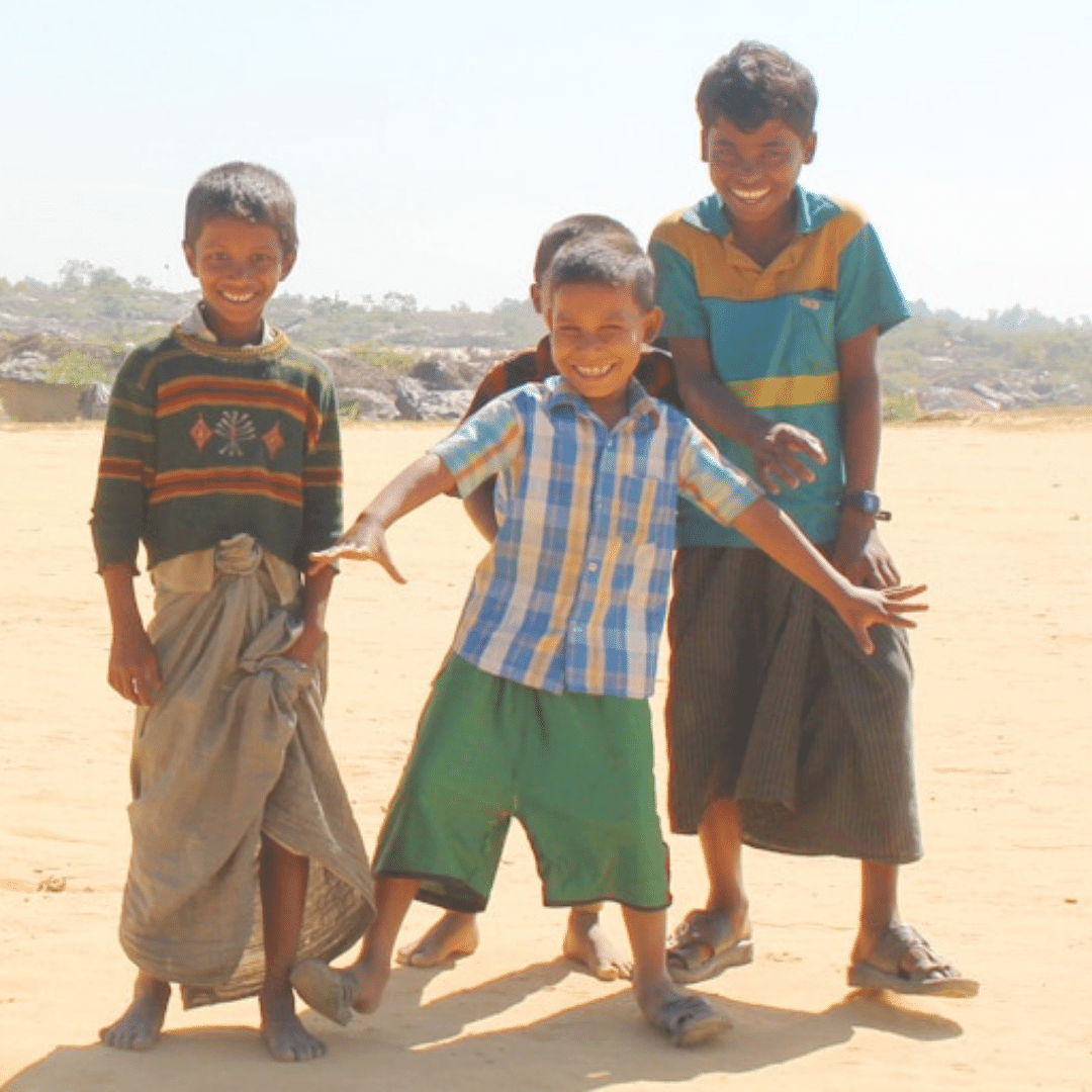Four boys around age 6 - 12 are standing outside on the dirt, they are all smiling at the camera. In the background is a large refugee camp in Bangladesh, where they live. Click on the image to read the blog.