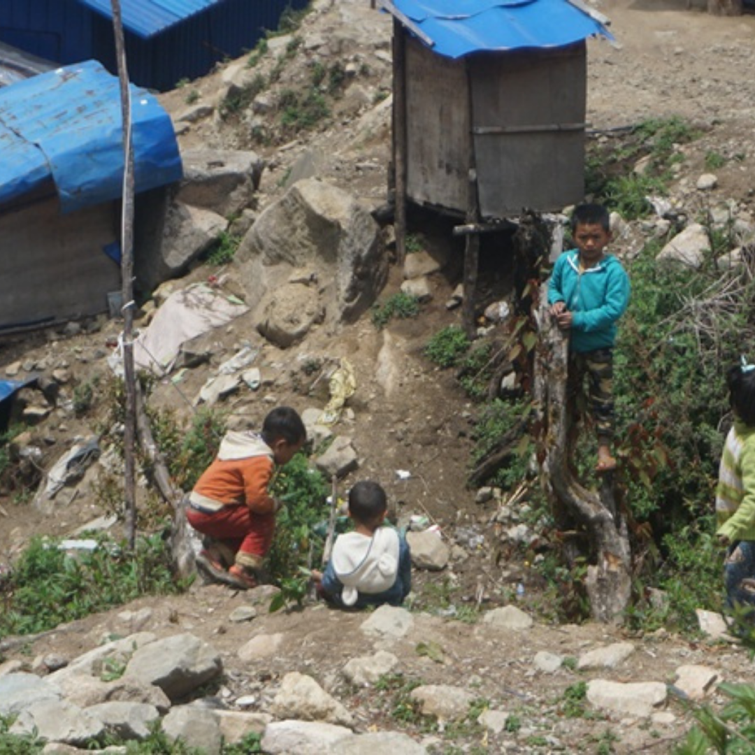 A remote mountain camp in Kachin State. Image shows three small boys playing on a hillside amongst the rocks. There is a small wooden hut beside them, which looks like a latrine. Other wooden structures are in the background, with blue metal roofs. One boy is standing up facing the camera and the other two boys, around the age of 5 are crouched down, facing away.  