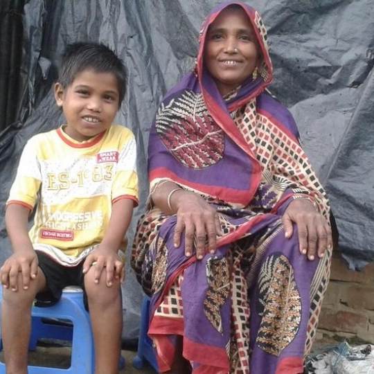 Narul, a young Rohingya boy. He is seven years old and dressed in a yellow tshirt. He is sat on a small blue plastic stool with his hands on his knees. His mother is sat next to him dressed in a purple patterned Sari and headscarf. She is also smiling. You can click on the image to read the blog post 