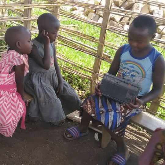 three Congolese refugee children sat inside a wooden shelter on wooden benches. One child, an older boy of around 8 years old, is holding a grey radio on his lap. The other two girls are sat next to him listening intently to the radio. You can click on the image to read the blog post 