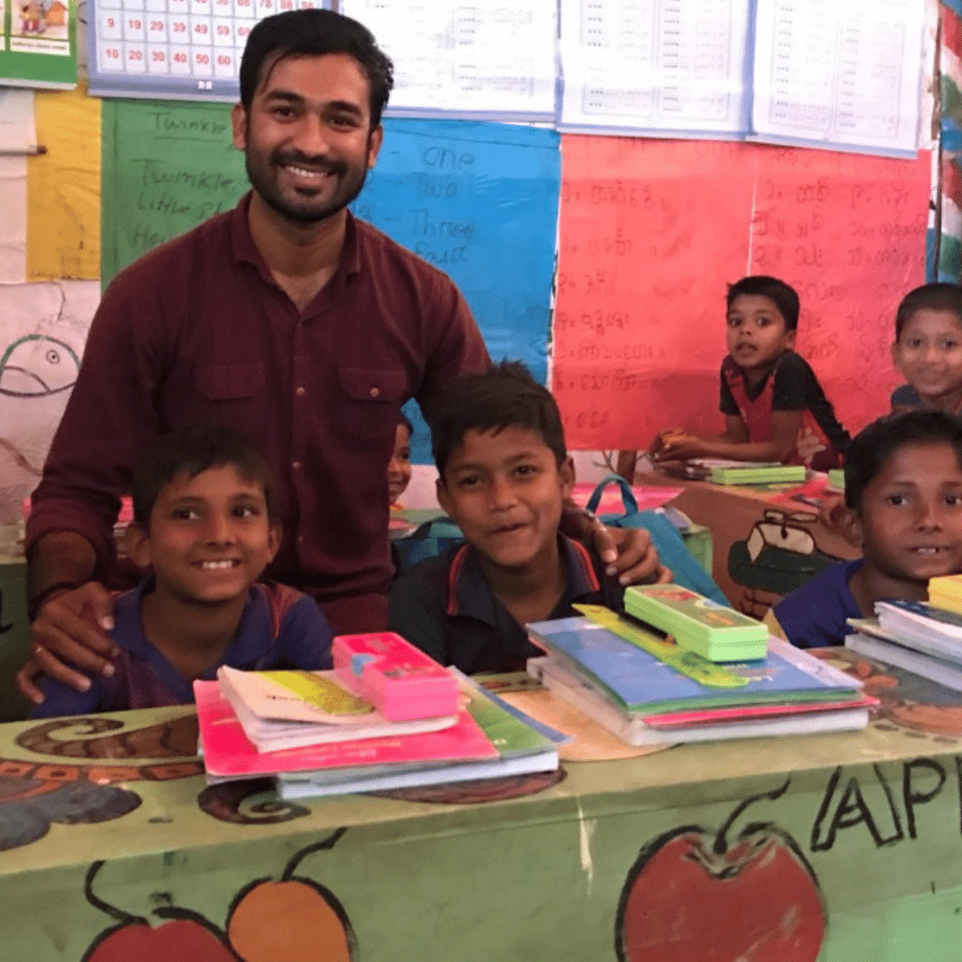 This is an image of Somorjit, a project officer in Bangladesh. He is kneeling behind two students in a colourful classroom and is smiling at the camera. Somorjit is wearing a maroon shirt and has his arms around the two students in front of him. The students are two boys who are also smiling and are sat behind a colourful table with books and stationary piled in front of them. You can click on the image to read the blog post. 