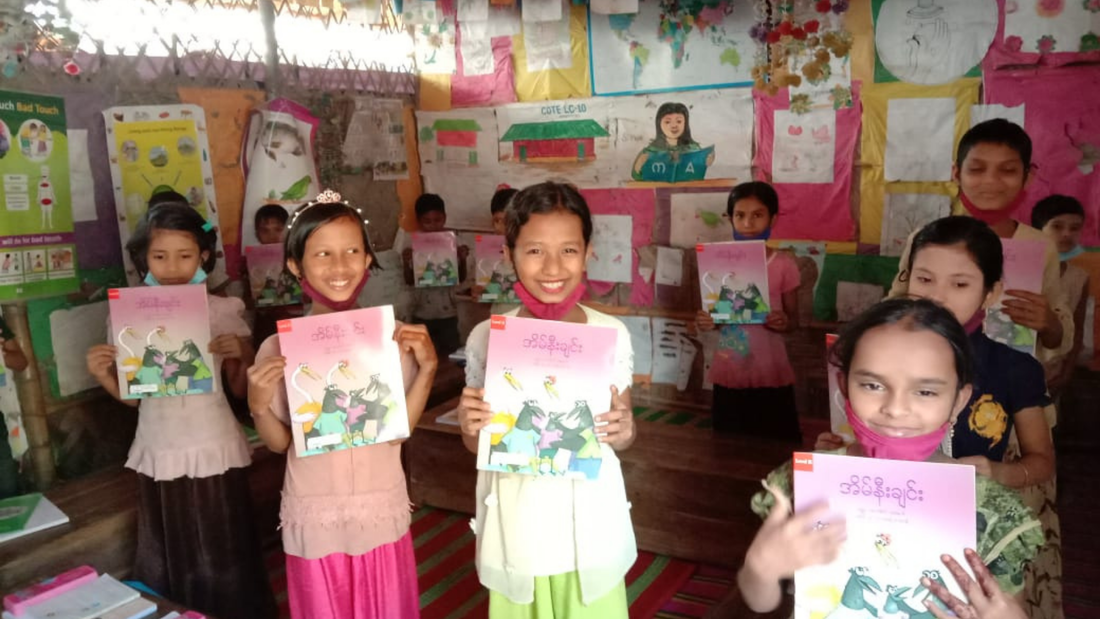 Rohingya children stood with new textbooks in their camp classroom. They are all smiling. 