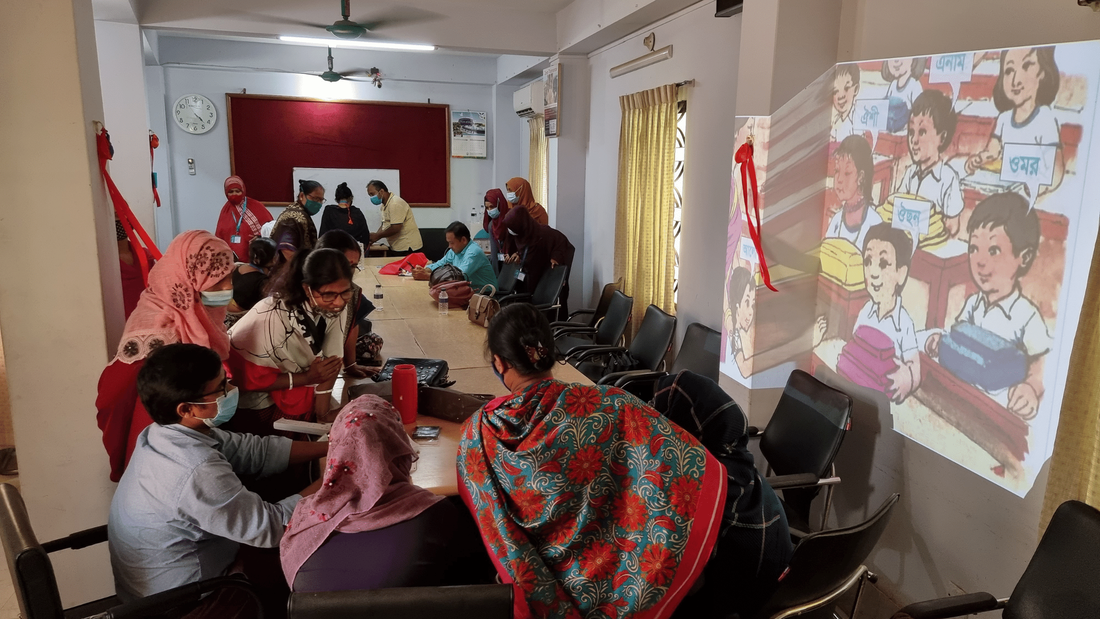 A group of Bangladeshi teachers taking part in a training session on how to use digital equipment. They are in a white room, with a projected screen on the wall showing a cartoon of a classroom. 