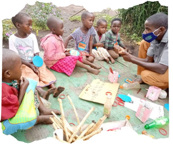 Group of young Congolese children sat on a mat outside with a teacher and homemade learning materials. He is explaining counting to them with some sticks. 