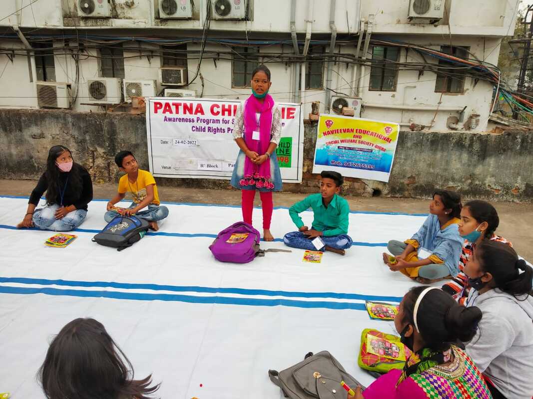 A group of Indian children are sat on a white and blue mat of their rooftop classroom. Many have backpacks and books in front of them. One girl wearing a bright pink scarf is stood up talking. 