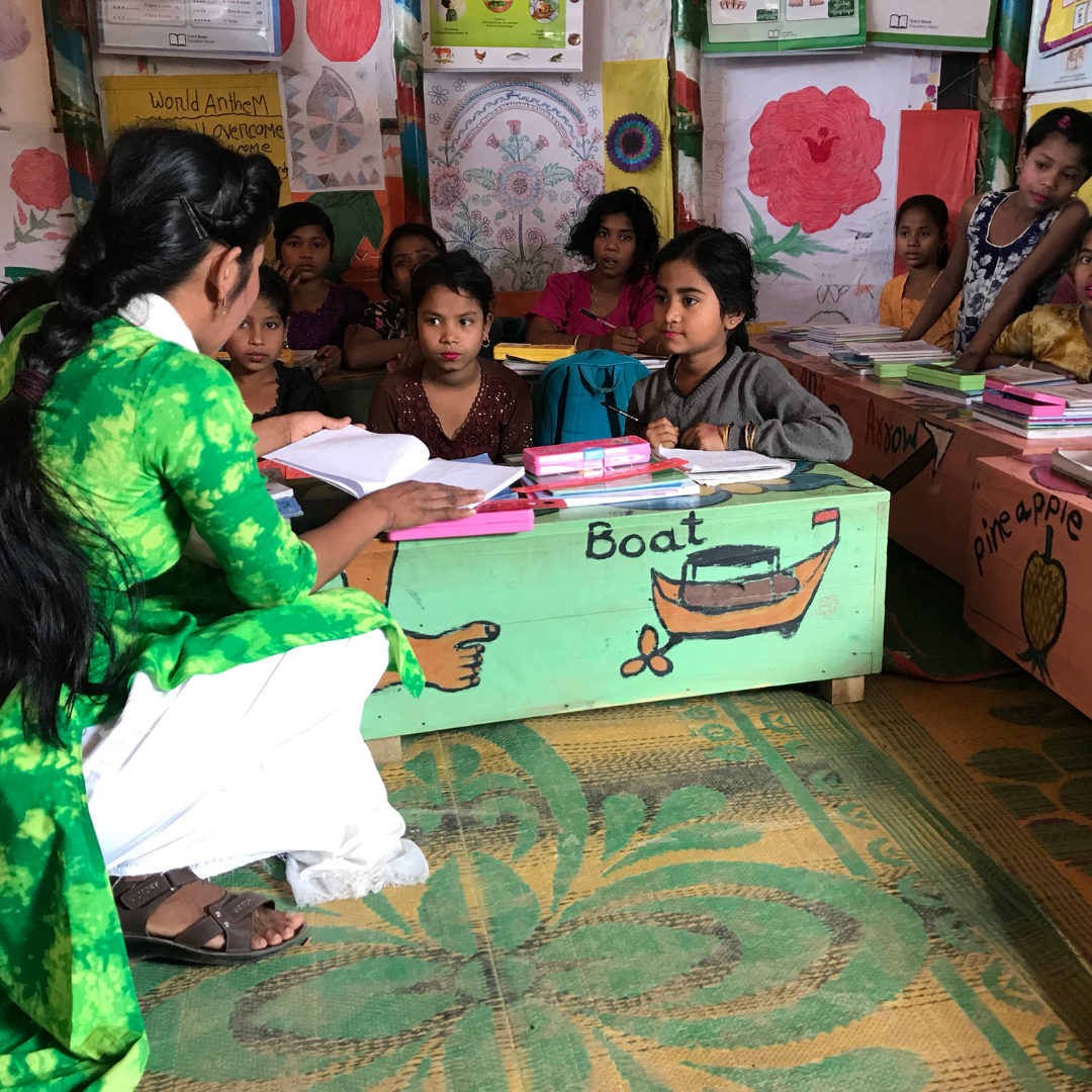 A female teacher sat on the floor in a small classroom. She is talking to a young girl at the front of the class who is sat on the floor at a low desk with another two girls. Five other children are sat at tables behind them. The classroom is bright and colourful, with childrens' pictures lining the walls. Click on the image to read about teaching in a mega camp