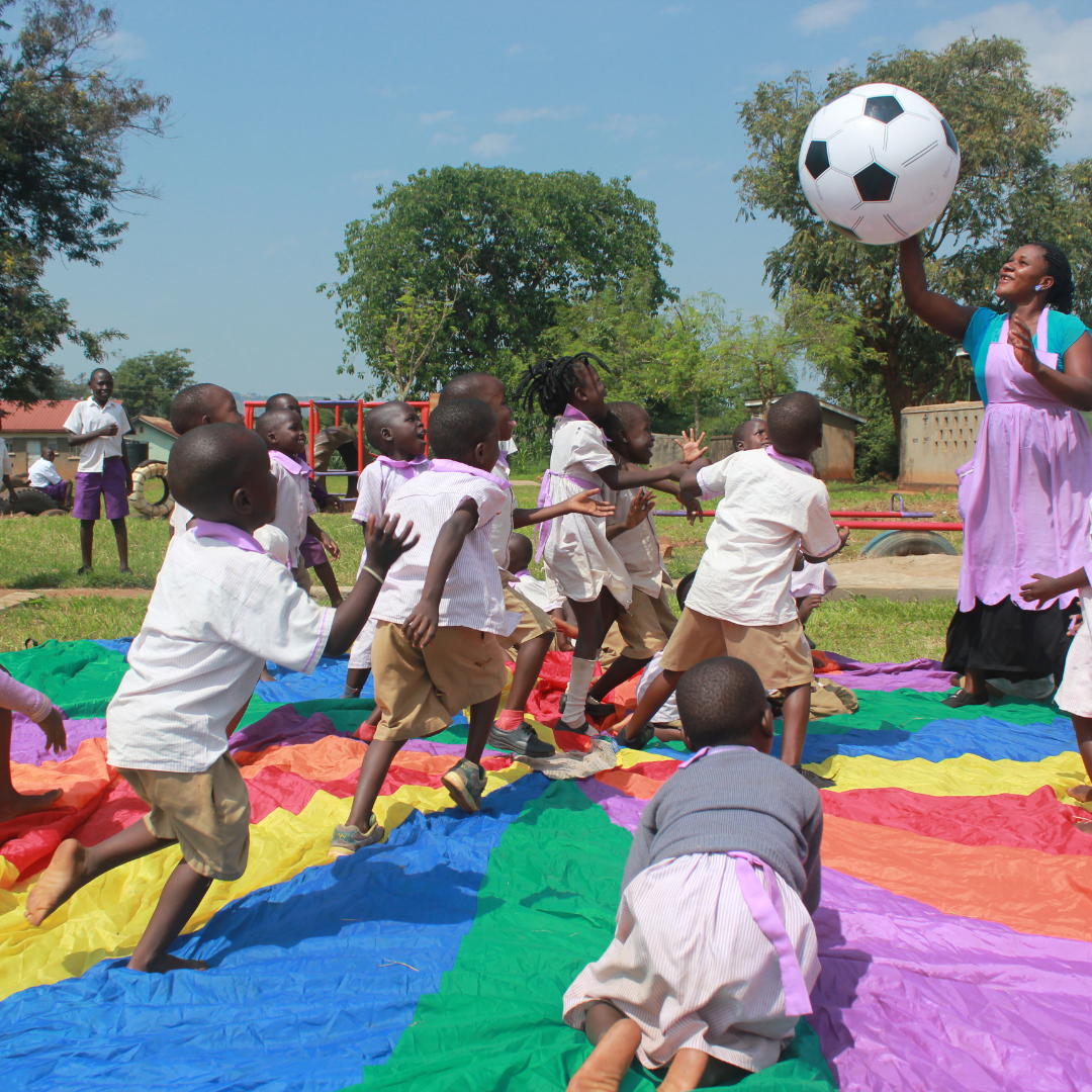 Image shows a small group of Uganda pre-school children running towards a large football being held up by a female teacher who is about to throw it to them. They are standing on a rainbow coloured parachute laying on the floor. The children are in a playground and there is play equipment and trees in the background.