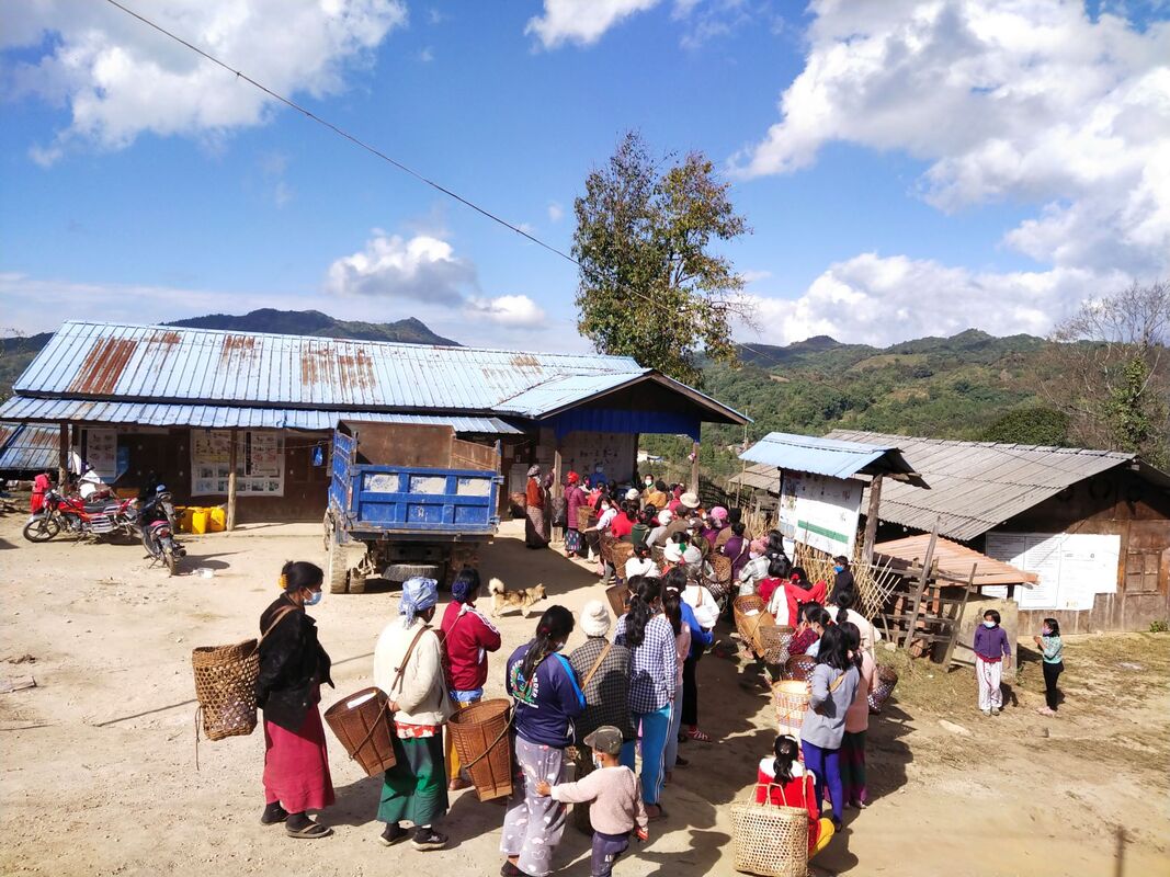 Kachin people queuing up outside a wooden building to register for humanitarian assistance. A desk at the front of the queue is staffed by someone in full PPE. Everyone is wearing masks. 