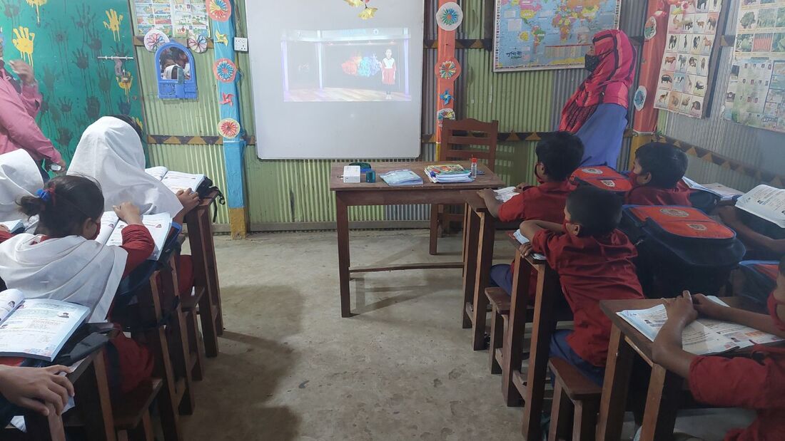 A classroom in Bangladesh full of children in uniform are watching a video on a projector screen at the front of class.
