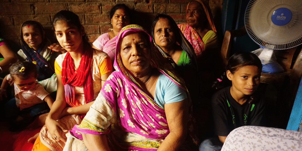 A group of Indian women, dressed in bright coloured clothes and headscarves. They are sat down in a dark brick room with a fan behind them. The women 5 women are looking at the camera with serious faces. Two children and a baby are sat with them, looking at the camera and smiling. 