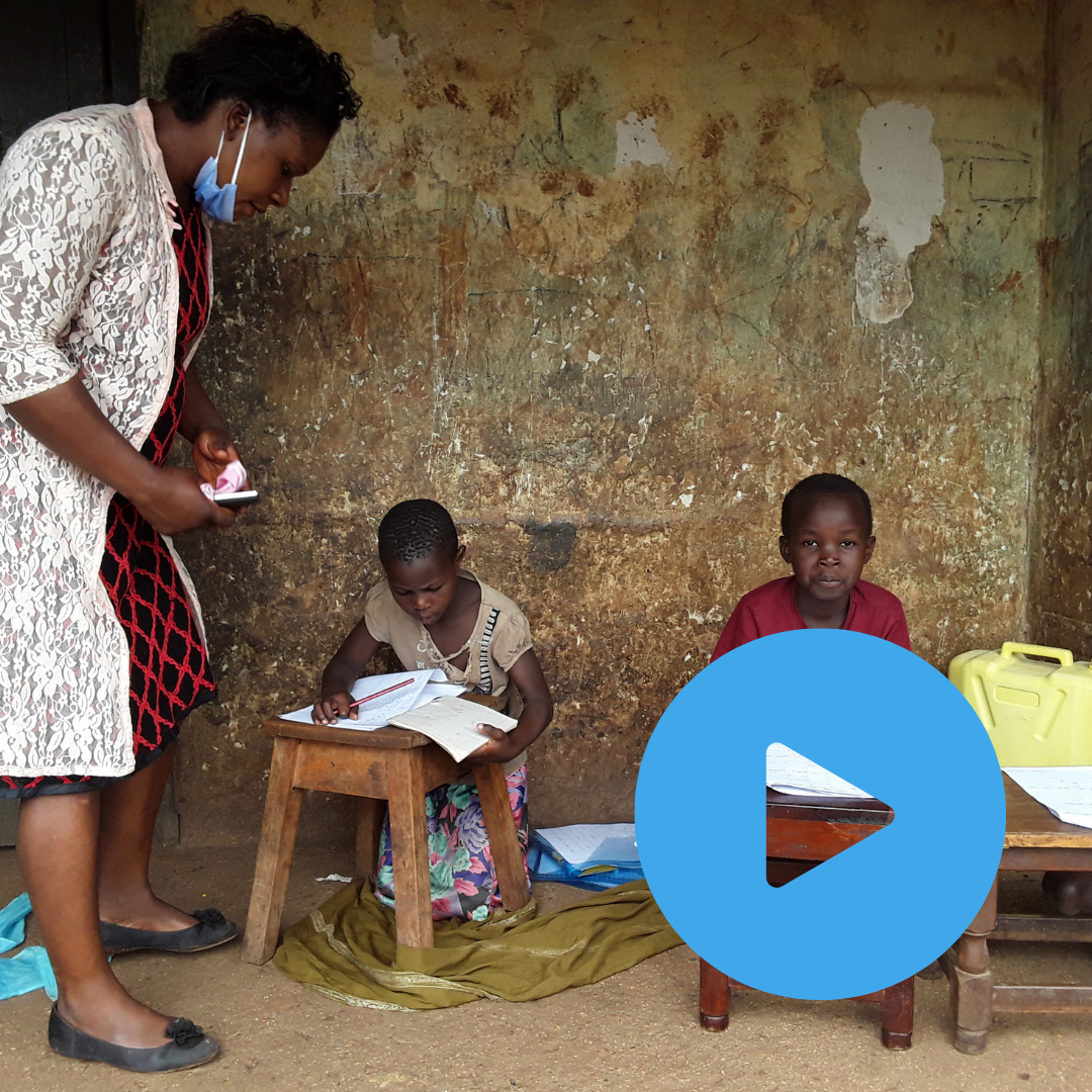 Ugandan teacher visiting two young children at home to deliver homework packs and check in on them. The two children are sat at little stools doing work as the teacher stands over them. You can click on the image to read the blog post. 